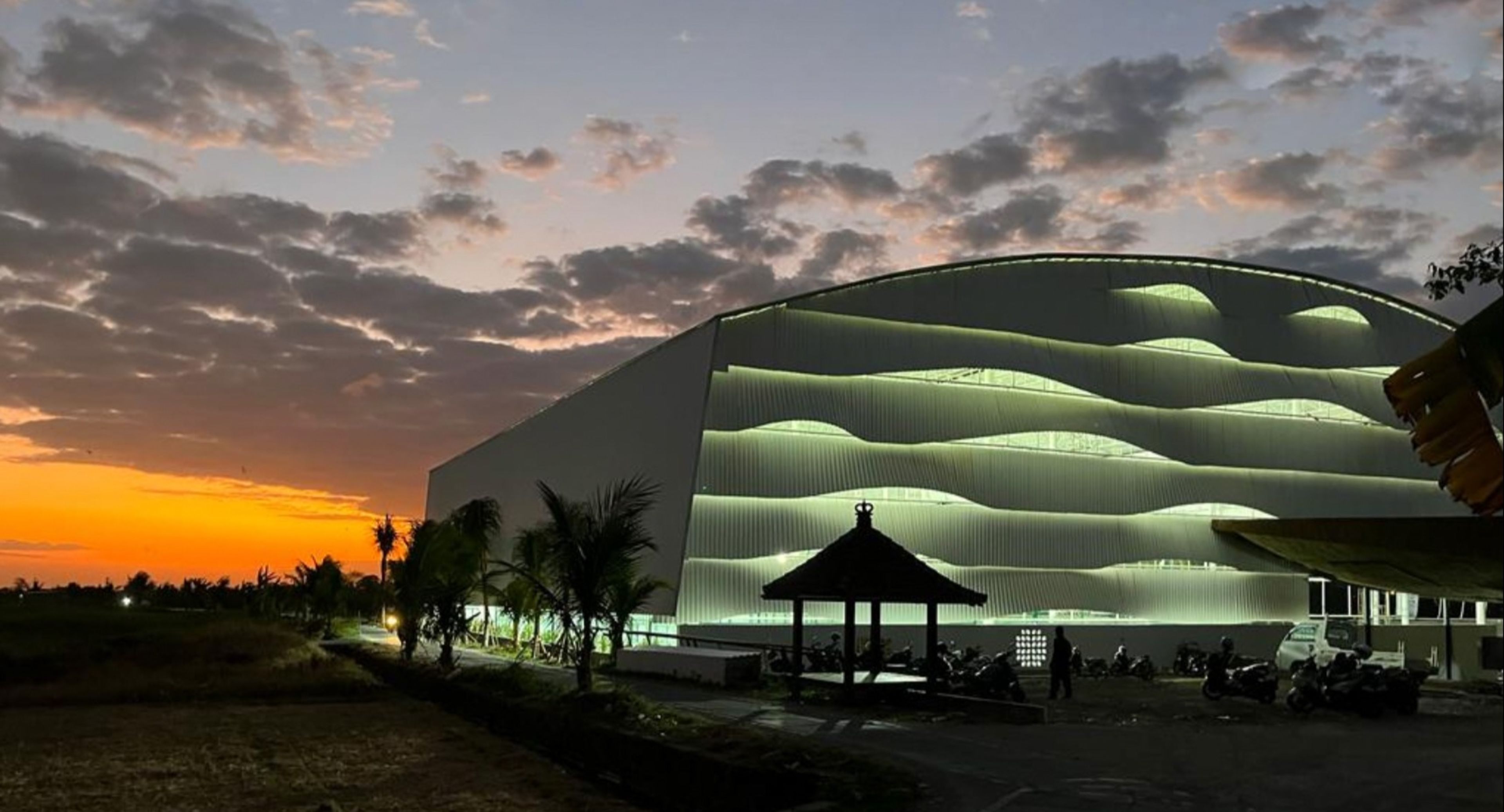 Bali Padel Academy exterior at sunset in Canggu showing the distinctive wave-shaped roof facade, tropical landscaping, and warm golden light