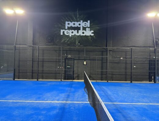 Floodlit indoor padel court with blue turf and branded back wall at Padel Republic Wallasey